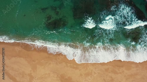 Aerial drone shot of turquoise ocean waves rolling onto sandy beach shoreline. Scenic coastal landscape with sea foam and clear water texture from above