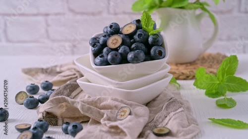 A studio shot of a white bowl stacked on rustic linen, overflowing with fresh blueberries and a mint garnish. Soft, natural light illuminates the scene against a light brick wall, creating an airy atm