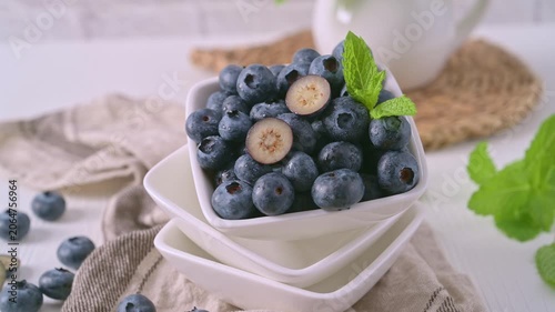 A close-up studio shot of a stack of white square bowls filled with fresh, plump blueberries and a sprig of mint. The composition features soft, bright lighting, rustic linen, and a neutral background