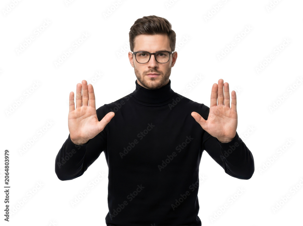 Fototapeta premium A young man in a black turtleneck sweater and glasses holds up his hands in a stop gesture on a white background.