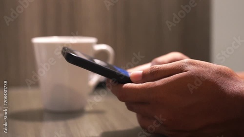 Man drinking hot coffee from white mug sitting at the table and typing text holding mobile phone in the hand. Closeup relax lifestyle video