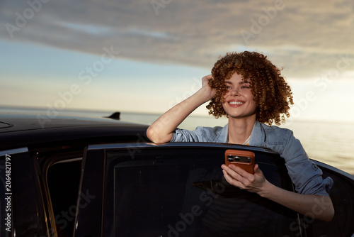 Fotografija Cheerful woman smiles from a car window at sunset, casual denim jacket, holding