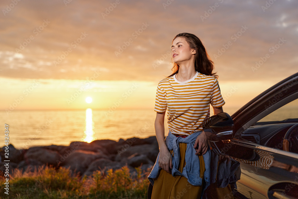 Fototapeta premium A young woman stands beside a car at sunset, wearing a striped shirt and denim jacket tied around her waist, looking toward the glowing horizon and calm coastline.