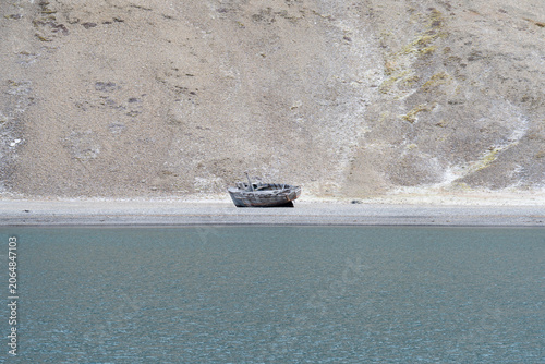 Fotografía Abandoned wooden boat beached on a pebbly Arctic shore, reflected in calm waters beneath towering barren mountains, conveying solitude and decay