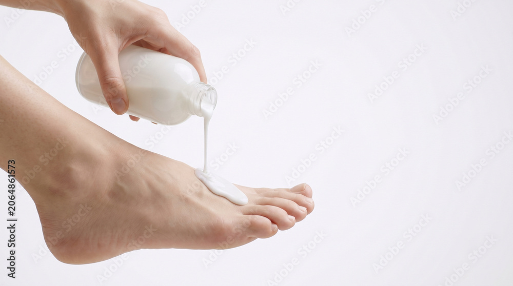 Naklejka premium Close up of hand pouring white milky lotion from minimalist bottle onto top of foot for skincare hydration