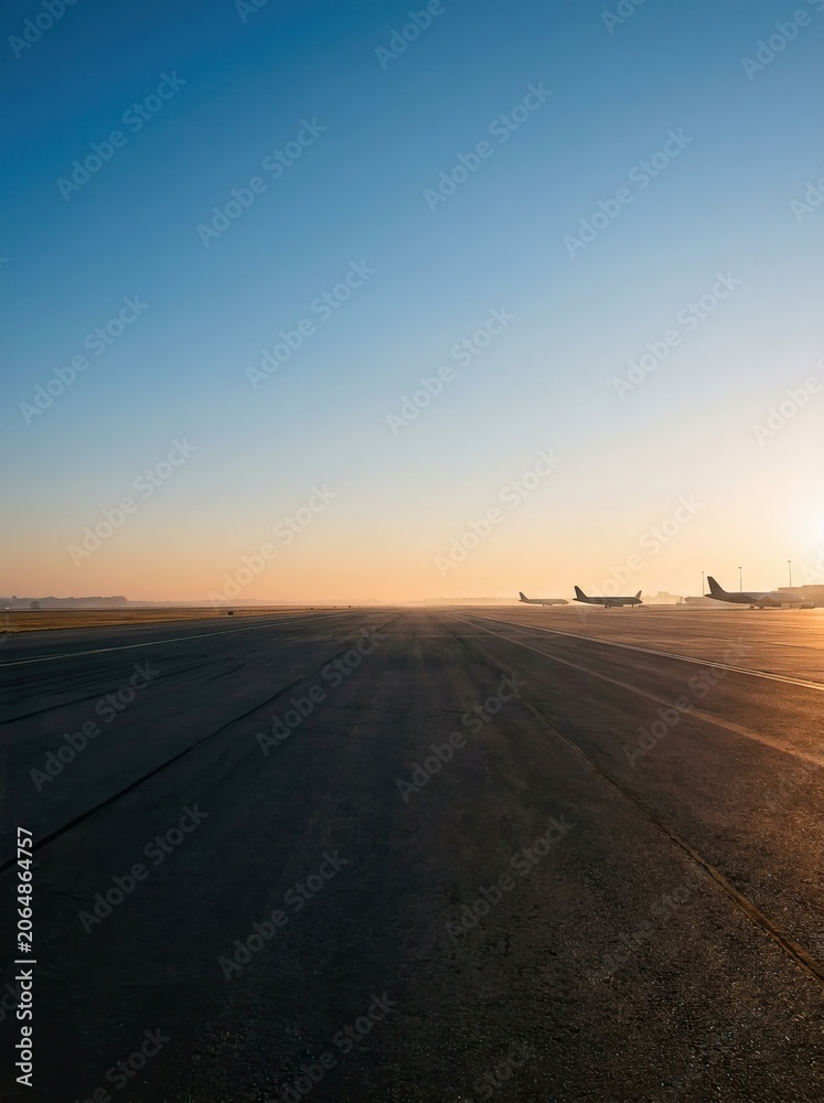 Fototapeta premium Two airplanes take off from a long airport runway. The sun rises in the distance, casting a warm glow on the tarmac.