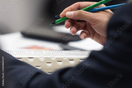 A man's hand dressed in a suit holds two colored pencils, one blue and one green, ready to underline religious scriptures. The scriptures are open, featuring thumb index tabs.