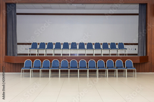 In an empty room, there are two arranged rows of chairs. One row is located on a pulpit, the other below on the floor. The chairs are blue and beige.
