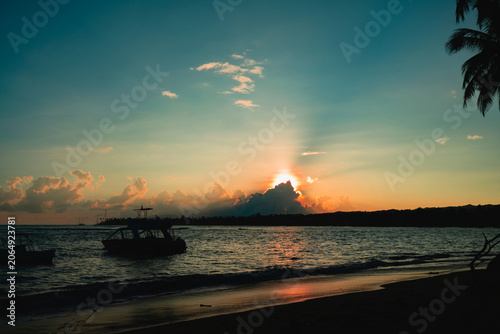 Dramatic tropical beach sunset with boat silhouette and sunburst.