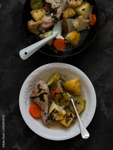 Stewed chicken with potatoes, zucchini, cauliflower and herbs in a cast-iron skillet is a comfortable healthy meal on a dark background, top view