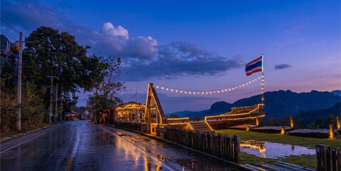 Scenic rural road at nightfall decorated with glowing string lights, dark blue twilight sky background with puddle reflection and copy space.