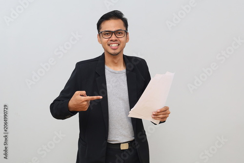 A successful young man, a businessman, points to an accounting document, financial report, or work document he is holding, standing alone on a white background.
