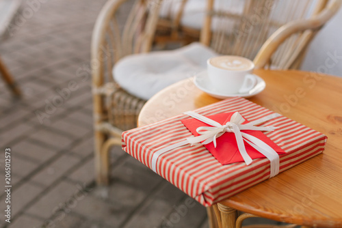 Close-up of a striped wrapped gift with a red envelope on a wooden table next to a blurred coffee cup.
