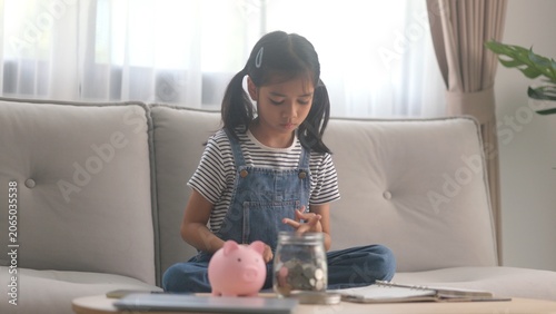 Smart female child counting saved coins next to glass jar on table. Cute little girl practicing basic economics and budget deposit planning inside cozy living room. Essential lifestyle content.