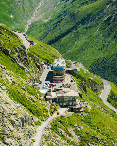 Obergoms VS, Switzerland - 10 August 2024: Aerial view of the historic Hotel Belvedere on the winding Furka Pass road surrounded by steep green mountain slopes and rocky terrain.