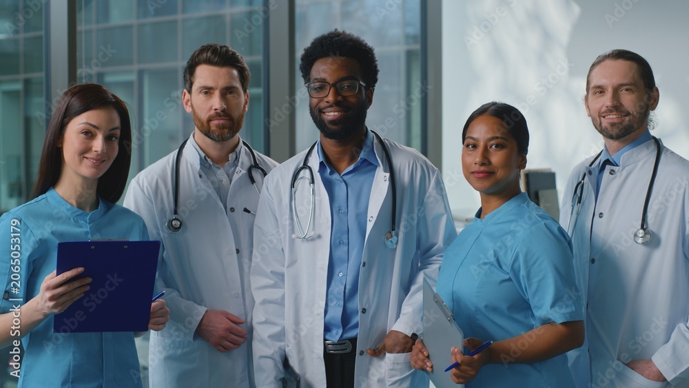 Fototapeta premium Happy smiling multiethnic doctors look in digital tablet talking discussing online health test results in clinic. Joyful team five surgeons looking at camera smile talk medicine teamwork in hospital