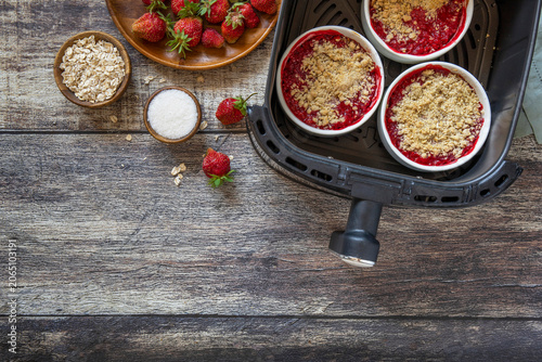 Homemade dessert traditional English crumble cake with strawberry in kitchen gadget modern air fryer on wooden background. Lifestyle trendy of new cooking. Top view flat lay background. Copy space.