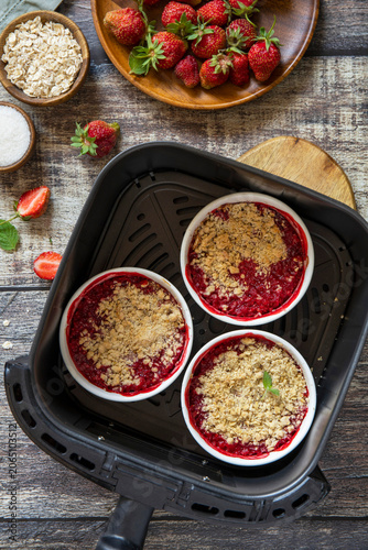 Homemade dessert traditional English crumble cake with strawberry in kitchen gadget modern air fryer on wooden background. Lifestyle trendy of new cooking. Top view flat lay background.