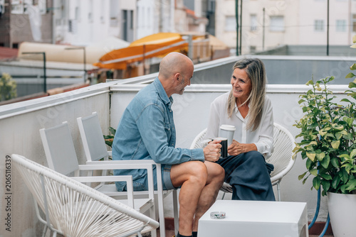 Two caucasian middle-aged adults share a joyful moment over coffee on a sunlit outdoor terrace, radiating warmth, connection, and relaxed lifestyle