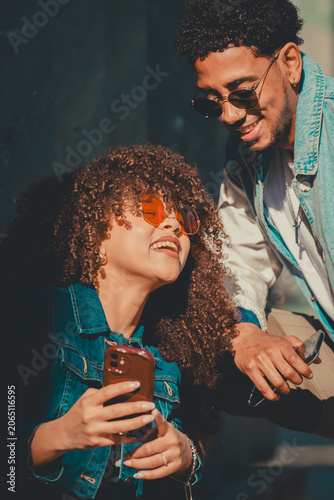 Young stylish couple sharing a fun moment looking at a smartphone outdoors, smiling in natural sunlight against a teal wall