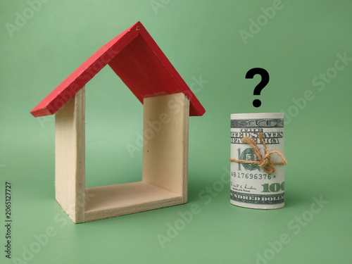 A miniature wooden house frame stands next to a rolled dollar bundle beneath an overlaid black question mark
