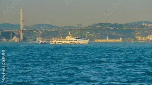 A white passenger ferry cruises through the blue waters of the Bosphorus in Istanbul, featuring a large suspension bridge and hilly cityscape under soft daylight