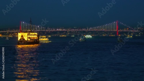A beautiful night scene in Istanbul featuring glowing passenger boats cruising on the Bosphorus with a brightly lit suspension bridge spanning the dark water