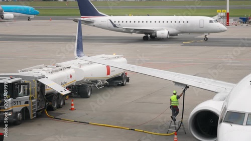 Jet fuel filling process of passenger airplane on airport apron with another commercial airliner taxiing in the background for aviation transport industry traffic logistics service operations.