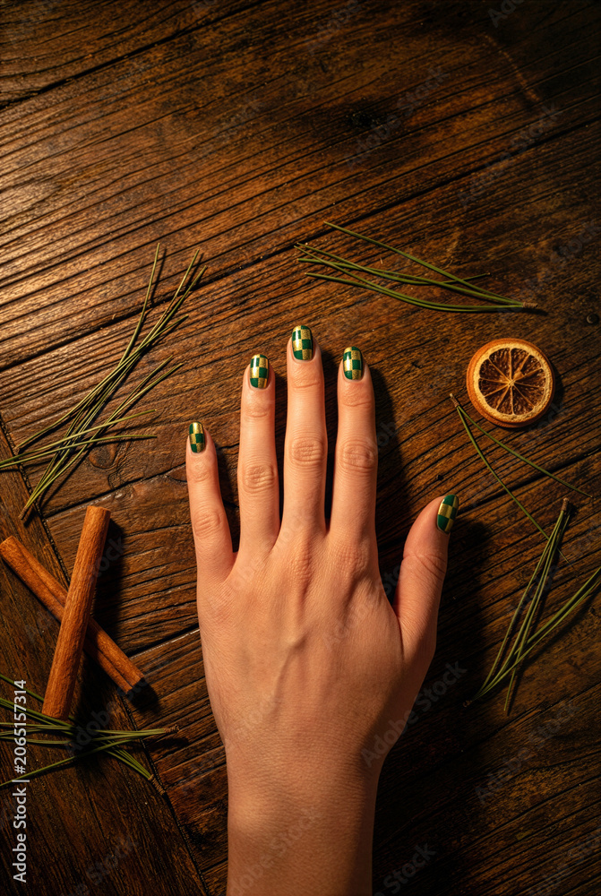Fototapeta premium Female hand displaying trendy green and gold checkered nail art on a rustic wooden table with pine needles, cinnamon, and dried orange.