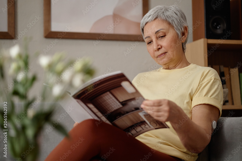 © TrueFrame Collective - Senior Asian woman reading magazine while sitting at home, showing glucose sensor on arm and independent diabetes management. Useful for healthcare campaigns, patient education