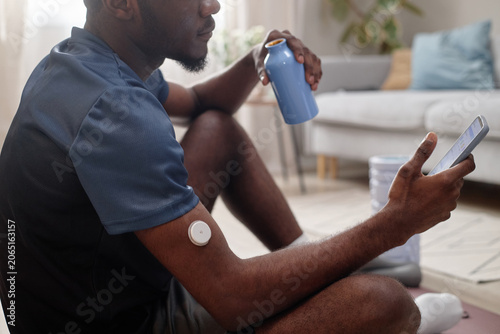 African American man checking smartphone fitness app while resting with water bottle, wearing glucose sensor after workout. Useful for diabetes tech, wellness, remote care marketing
