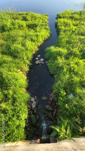 Dirty wastewater flows from drainage pipe through narrow channel toward river. Concept: sewage leak, environmental pollution and water contamination.