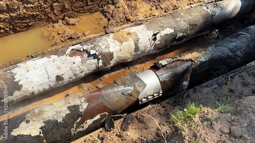 Old heating pipe leaks near temporary repair clamp in excavated trench. Concept: failed repair, worn pipeline, utility accident and emergency maintenance.