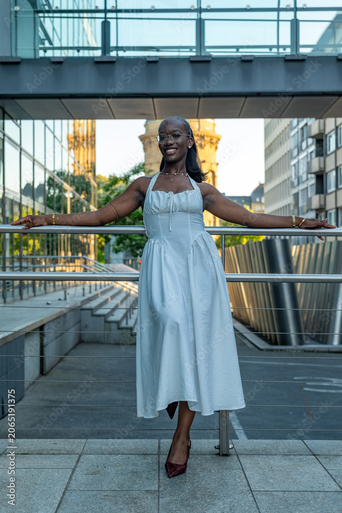 Fototapeta premium Woman stands on a railing wearing a white dress at a modern city location during sunset