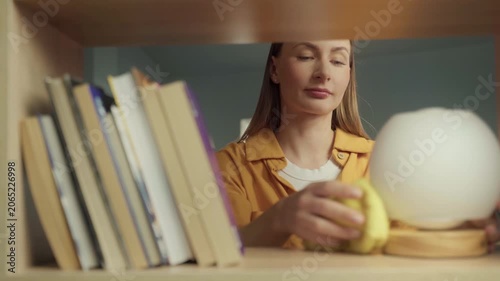 Young woman meticulously wiping dust from a wooden bookshelf and books with a yellow cloth, tidying up the living room during her daily cleaning routine and maintaining a neat home environment
