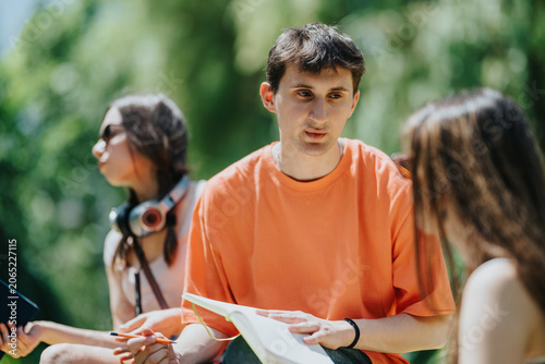A male student in an orange shirt studies and discusses notes with classmates outdoors. Three students sit together in a park exchanging ideas and reviewing a notebook.