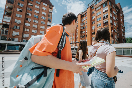 Students walking through an urban courtyard carrying backpacks and notebooks. A group of young people stroll between apartment-style buildings on a sunny day.