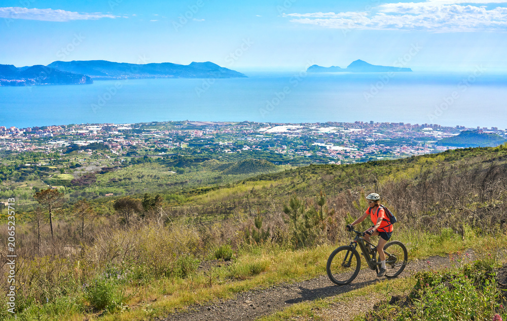 Fototapeta premium Smiling senior woman riding an electric mountain bike above Pompeii. Panoramic view of towns, mountains, and the Gulf of Naples, Active lifestyle, and healthy aging in Pompei, Campania, Italy.