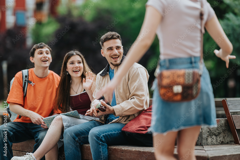 Obraz premium A group of university students sit on a bench smiling as they greet a friend approaching. The casually dressed friends chat and laugh together on an outdoor campus bench.