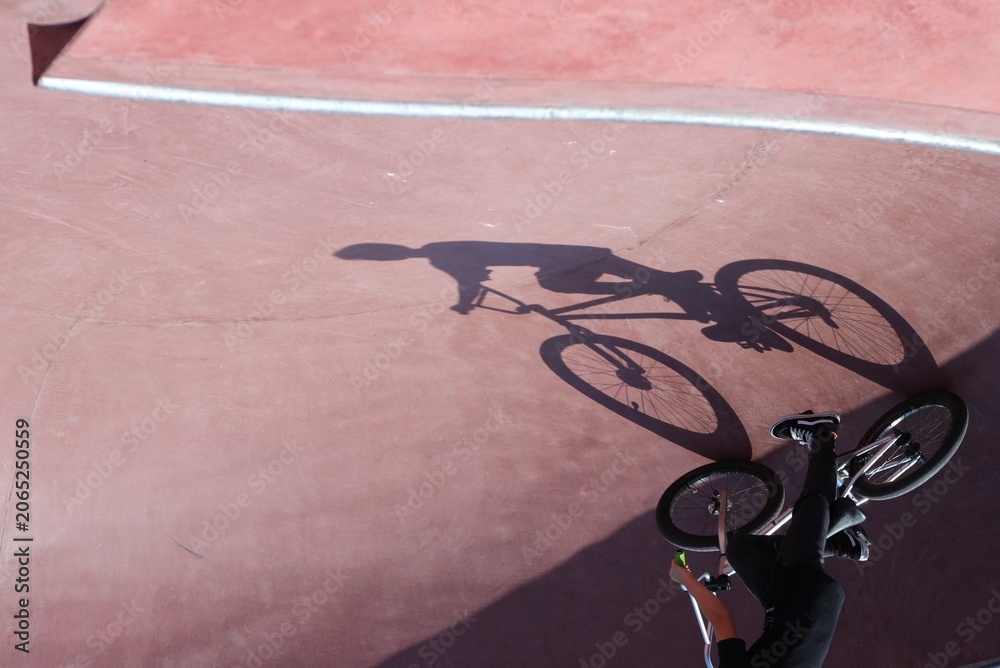 Naklejka premium A creative perspective shot showing the shadow of a cyclist on a red skate park bowl surface, with the rider visible in the corner.