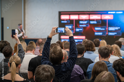 Audience captures presentation on smartphone during busy business conference, workshop slide visible on large screen.