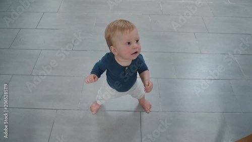 An innocent toddler tries to stand on his feet. A brave little child explores his abilities. A small child observes his home environment with interest. A child learns to stand on a tiled floor