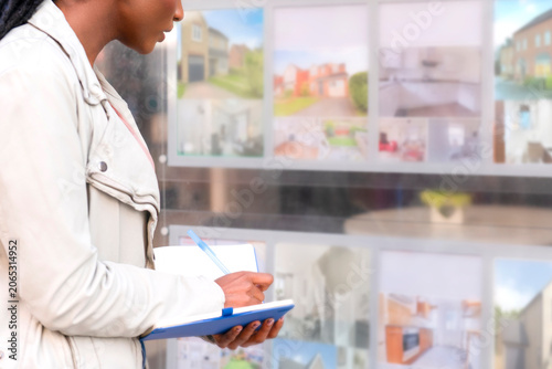 Woman writes in notebook while looking at property listings in real estate window display in a city