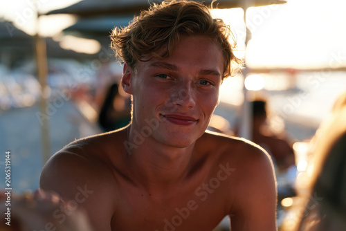 A young man with curly hair smiles toward the camera at the beach during sunset, capturing a joyful moment filled with warmth and relaxation by the ocean.
