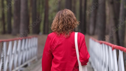 A young woman with curly hair in a red jacket walks through a spring park, relaxing after a working day.