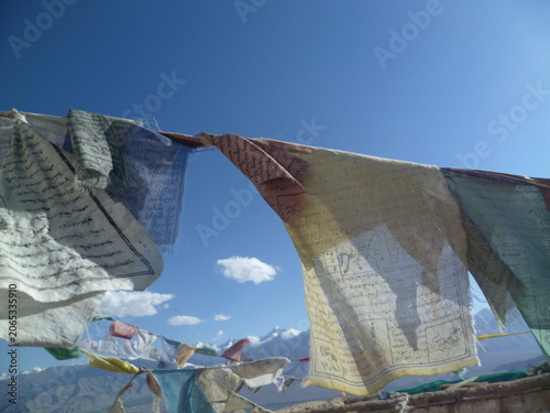 Traditional colorful Tibetan prayer flags fluttering in the wind against a deep blue sky and distant snow-capped mountains in Ladakh.