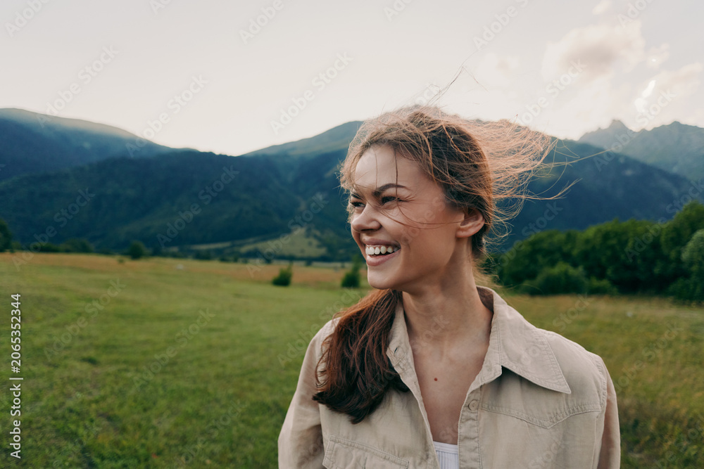 Obraz premium Smiling woman in casual shirt outdoors, bright mountains in the distance, open field under daytime sun, lifestyle portrait conveying happiness, freedom, and connection with nature.