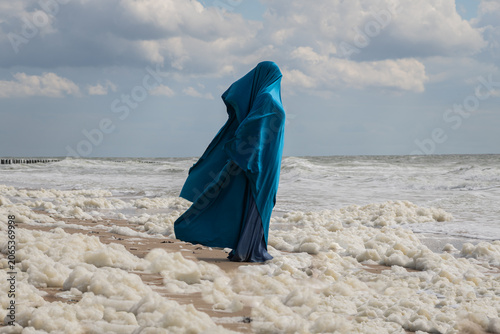 anonymous woman on the beach in blue dress and fabric standing in  foam of ocean