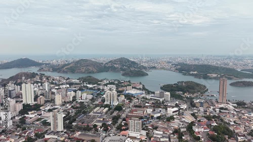 Panoramic aerial view of Vitória, Espírito Santo, Brazil, showcasing the urban skyline, bays, islands, hills, and coastal landscape beneath a dramatic cloudy sky.