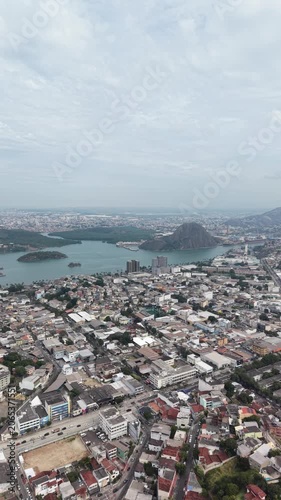 Panoramic aerial view of Vitória, Espírito Santo, Brazil, showcasing the urban skyline, bays, islands, hills, and coastal landscape beneath a dramatic cloudy sky.